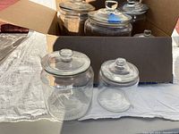 Photo of two clear glass apothecary jars placed on fabric with a cardboard box in the background containing more jars.