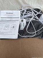 Grey plush Brookstone electric blanket with tangled white wired dual control units and printed instruction manual, displayed inside a cardboard box.