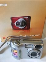 Front view of Nikon Coolpix 3100 camera with wrist strap and Canon PowerShot A3100 IS box in the background.