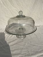 Full view of clear glass cake stand with dome lid on a white textured cloth background.