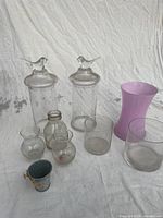 Photo shows assorted decorative vessels and containers arranged on white backdrop. Two tall clear glass jars with lids topped by glass bird figurines are centered at back. Multiple small clear glass vases and containers along with a light purple glass vase and metal bucket with floral painting are positioned in front.