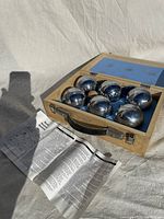 Closed open wooden carrying case containing six metal petanque boules with engraved lines, placed on a white fabric background with folded instruction sheet.