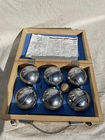 Open wooden carrying case showing six steel petanque boules, one wooden jack ball, and game instructions.