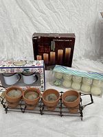 Lot overview showing used terracotta pot candles in metal holder, 2-piece cup set in floral box, 6 flameless pillar candles in box, and 10-pack white outdoor tealight candles in plastic packaging.