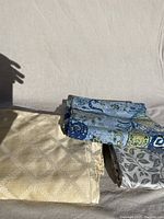 Three folded tablecloths arranged against a plain fabric backdrop, visible partial patterns of gold diamond and blue paisley designs