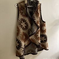 Full view of sleeveless wool vest hanging against a wall showing draped waterfall style front and raised circle pattern in tans, rusts, and dusty browns.