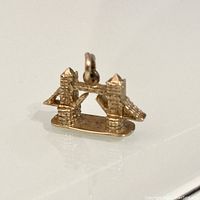 Front and angled view of a small 9 karat gold charm shaped like the London Tower Bridge, showing detailed towers and suspension cables.