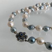 Close-up of round filigree clasp with seven pale blue sapphires and adjacent smoke-grey and cream baroque pearls.
