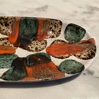 Close-up showing the drip glaze pattern in green, orange, and brown on off-white background with textured spots, details of glaze colors and texture.