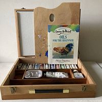 Open wooden box showing canvas board, paints, instructional book, and compartments with supplies