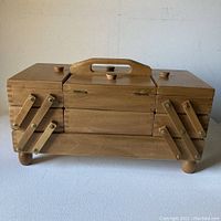 Front view of closed wooden sewing box showing handle, hinged lids and fold-out tiers.