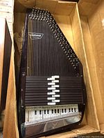Full view of trapezoidal autoharp in cardboard box showing body shape, strings, chord bars and logo