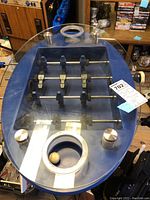 Top view of the blue oval foosball table surface showing seven blue player figurines on rods and a circular beer cooler with wooden ball inside.