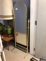 Full view of brass framed floor mirror beside wooden glass table and green glass lamp.