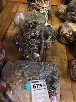 Glass vase filled with silver and black costume jewelry and multiple bags of assorted costume jewelry in the background on wooden surface