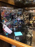 Six clear containers filled with assorted costume jewelry items on a wooden shelf.
