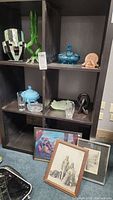 Black shelving unit with multiple glass and crystal decorative items including a green bud vase, blue lidded dish, clear crystal vase, and framed art leaning against it.