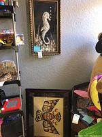 Wide view showing the framed seashell seahorse shadow box hanging on the wall above the Indigenous signed print leaning against the wall.