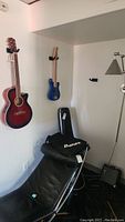 Wide view of room wall showing red sunburst acoustic guitar, blue electric guitar, black Ibanez soft case, hard case, black Fender amp and chair