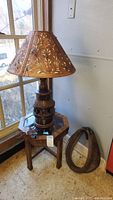Full view of wooden table lamp with floral carved leather lampshade, small side table with octagonal glass top, and pony collar on floor next to them.