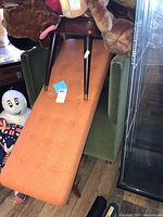 Orange button-tufted bench seat angled atop olive-green velour chair with brown stool legs visible