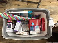 Top-down view of a plastic tub filled with craft supplies including boxed markers, holiday cards, wrapping paper roll, and sewing machine kit.