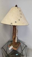 Full view of rustic table lamp showing cream shade with leather stitched trim and metallic conchos with base of intertwined wood branches and leather.