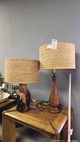 Two wood base table lamps with cork shades on a wooden side table with other household items visible in background.