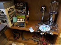 Photo of four toy slot machines displayed on a wooden shelf, including one smaller green slot machine bank with red lever in the foreground, a larger JUMBO SLOT metal slot machine bank in the box, and two other slot machines in the background. Also visible is a unique clock made out of spoons and forks on the upper shelf and a vintage horseshoe wreath.