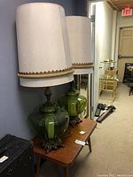 Pair of vintage green glass lamps with brass bases and large beige fabric shades on a vintage wooden table in hallway area.
