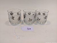 Front view of three clear glass tumblers with silver foliage designs and banding.