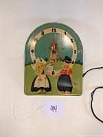 Front view of vintage Art Deco wall clock featuring Dutch boy and girl, windmill background, with green frame and red hands.