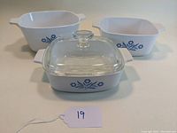 Three square white casserole dishes with blue cornflower design shown from front angle on flat surface, one with glass lid.