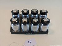 Front view of the full spice rack with eight metal shakers with black lids, all shiny aluminum bodies, sitting in a black metal rack labeled SPICES