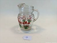 Front-side view of a clear glass pitcher with red tulips painted on its body, showing handle and pouring spout with notch.