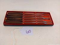 Six teak handled fondue forks arranged in original red box, showing colored tips and fork prongs.