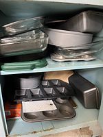Photo showing assorted glass casserole dishes, metal loaf pans, muffin tins including 6-cup and 12-cup sizes, with some trays stacked on a shelf.
