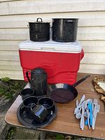 Red Coleman cooler with two large black metal pots on top, black metal coffee pot, four black metal cups, black frying pan, assorted white and blue reusable plastic cutlery on a wooden table near an outside wall.