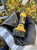 DeWalt cordless cutout saw placed on top of a worn Irwin electricians tool pouch outdoors on leafy ground, front angle view showing saw body and battery pack.