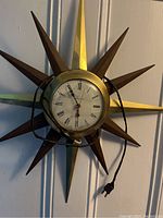 Vintage starburst wall clock with alternating wood and brass spokes, broken glass face showing Roman numerals, electric cord with two-prong plug.