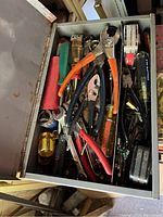 Open metal toolbox filled with assorted vintage hand tools including pliers and screwdrivers with various colored handles.