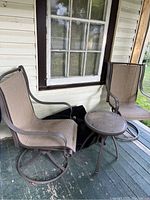 Two beige mesh swivel chairs with brown metal frames and a small round glass-top side table on a porch floor near a window.