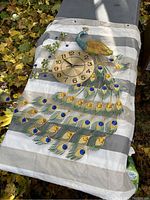 Photo of a metal peacock wall clock with detailed multicolored feathers and a round clock face, placed on a striped fabric near outdoor autumn leaves.