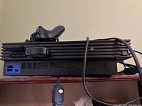Front view of PS2 console on shelf with controller and IR receiver on top, power cable hanging in front.