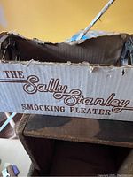 Cardboard box with 'The Sally Stanley Smocking Pleater' printed in vintage style font, showing significant wear and tears.