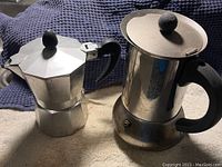 Two stove top espresso pots placed on a surface, one aluminum with octagonal shape and one stainless steel with round shape and spout.