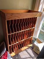Front and side view of the wooden mail sorting cabinet showing multiple vertical mail slots and wood grain finish, placed near a window with sunlight.