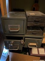 Three vintage metal filing units stacked together: one single index card drawer on left, one double index card drawer on right, and a small four-drawer unit below missing one drawer.