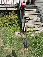 Front view of the red Yard Machines Y725 gas-powered weed trimmer standing on grass near wooden steps.