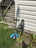 Both corded tools standing against house exterior showing trimmer with blue guard and hedge clipper with black and yellow handle.